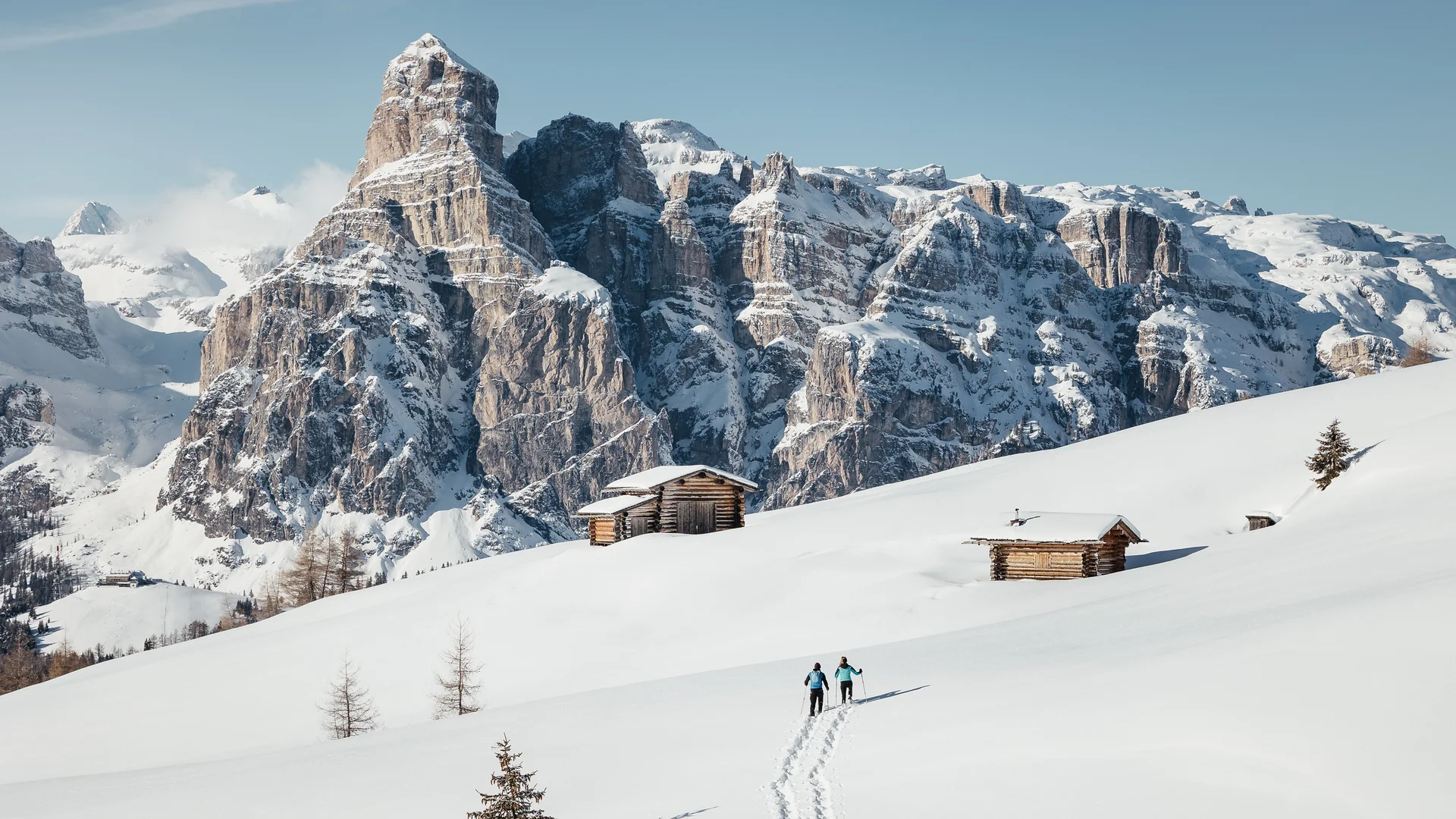 Zuhause auf Zeit im Hotel in Colfosco: Nature Hotel Delta Zwei Skifahrer auf verschneiter Alm mit Holzhütten vor schneebedeckten Bergen