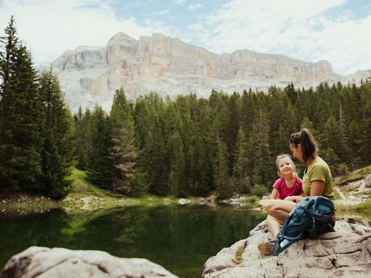 Zuhause auf Zeit im Hotel in Colfosco: Nature Hotel Delta Mutter und Tochter sitzen auf Felsen am See vor Wald und Bergen